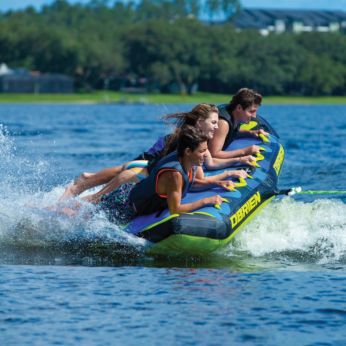 Three people enjoying an OBrien Bat Wing 3 Boat Tube on a lake, creating splashes.