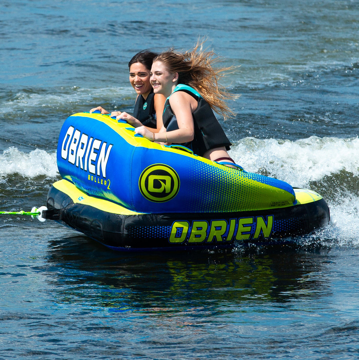 Two smile riding the OBrien Baller 2 Towable Tube, a blue & green inflatable.