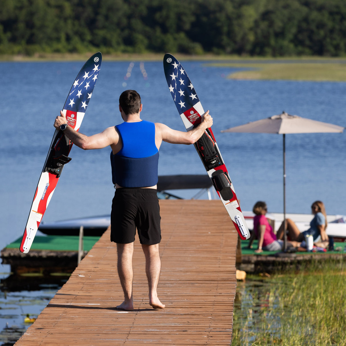 A man in a blue life vest walks barefoot on a dock holding the O'Brien Celebrity 68" Combo Waterskis with a flag design.