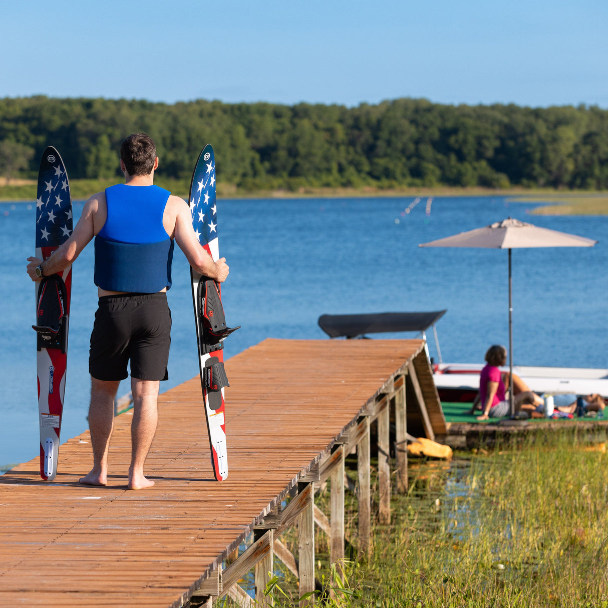 A man in a blue life vest stands on a dock holding the O'Brien Celebrity 68" Combo Waterskis, facing the water.