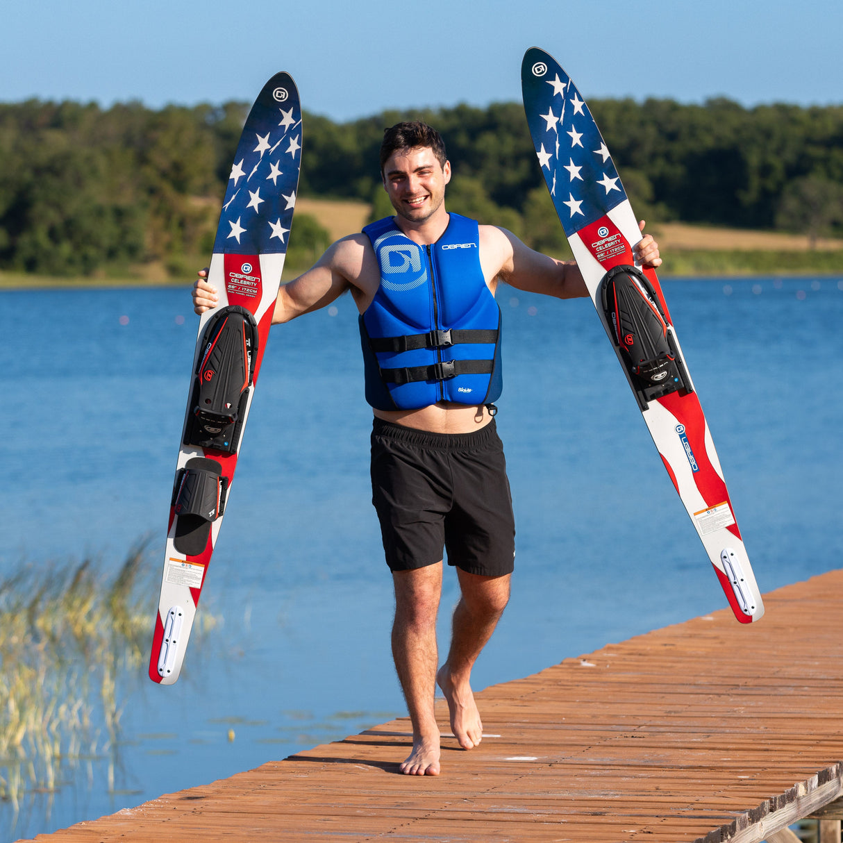 A smiling man in a blue life vest walks on a dock holding the O'Brien Celebrity 68" Combo Waterskis with a flag design.