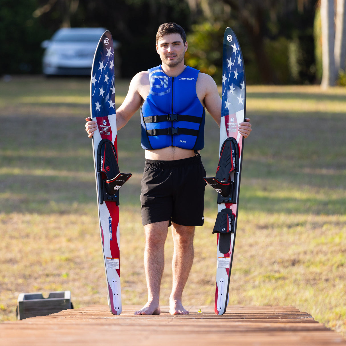 A man in a blue life vest stands on a dock holding the O'Brien Celebrity 68" Combo Waterskis, with a grass in the background.