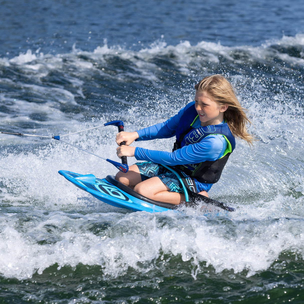 A young rider wearing a blue life vest kneeboarding on the O'Brien Radica, gripping a handle while carving through the water.