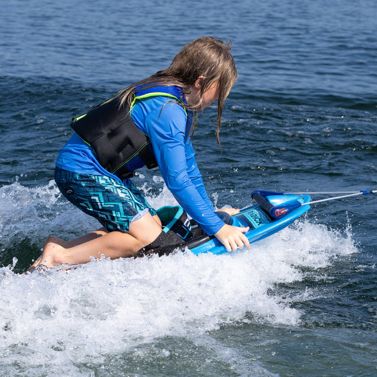 A young rider in a blue life vest kneels on the O'Brien Radica, adjusting position while gliding on the water’s surface.