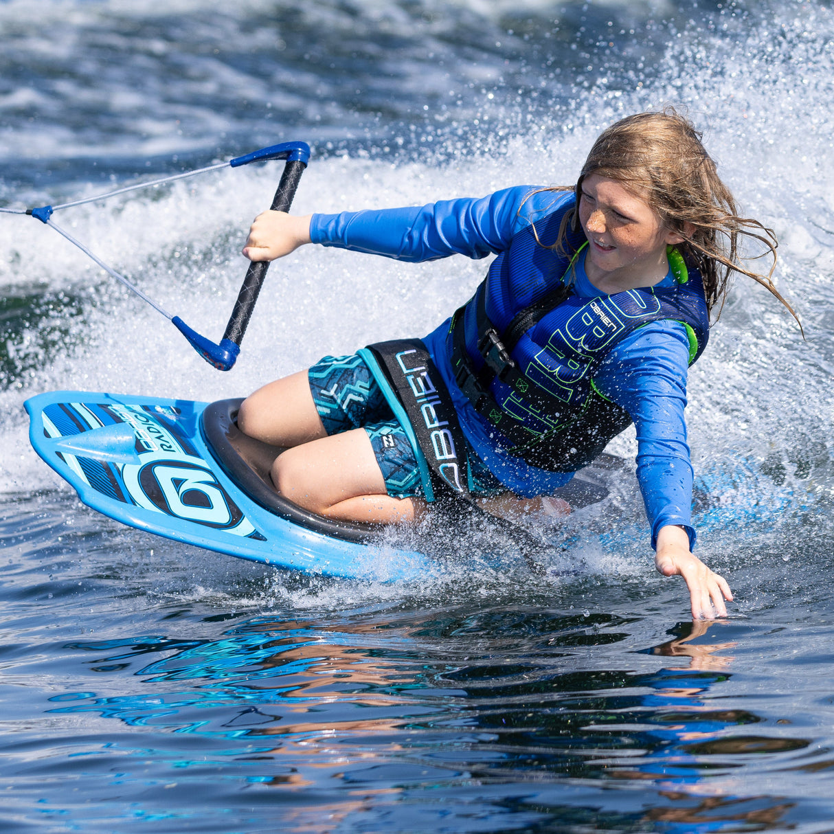 A young rider in a blue vest rides the O'Brien Radica kneeboard, cutting through the wake with a focused expression.