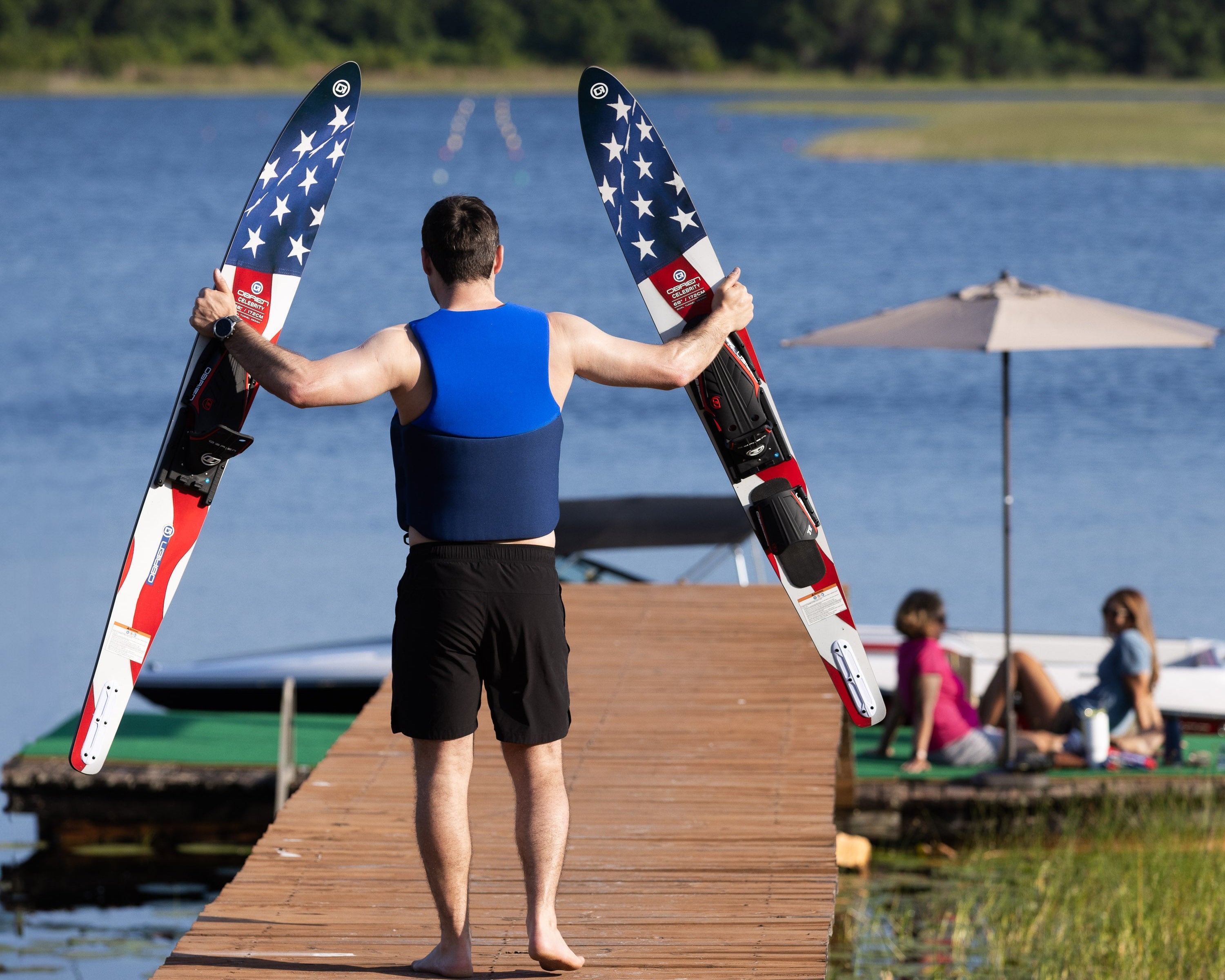A man in a blue life vest walks barefoot on a dock holding the O'Brien Celebrity 68" Combo Waterskis with a flag design.