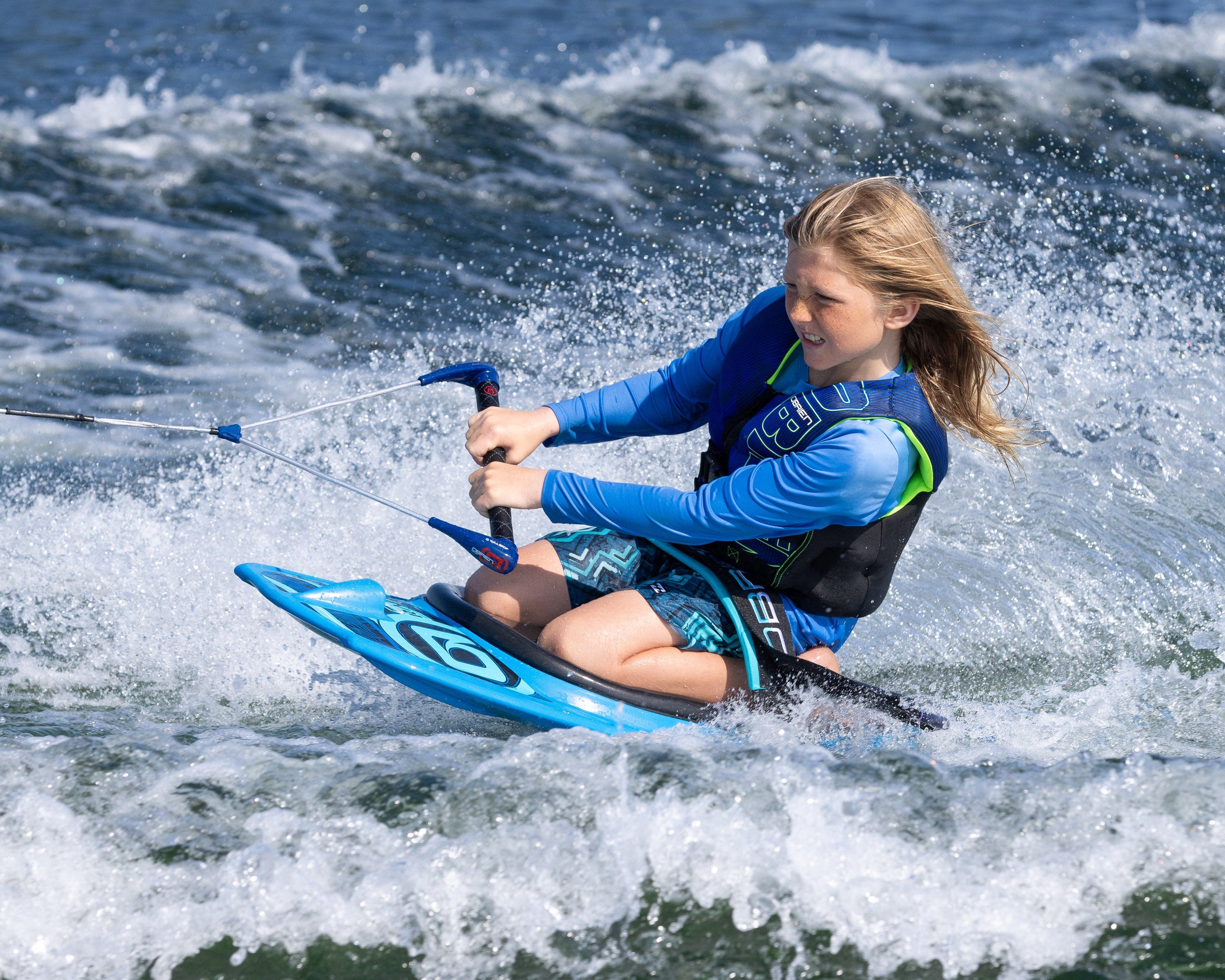 A young rider wearing a blue life vest kneeboarding on the O'Brien Radica, gripping a handle while carving through the water.